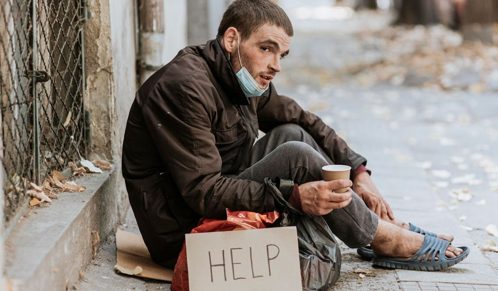 A man sitting outdoors holding a cup beside a sign that says Help